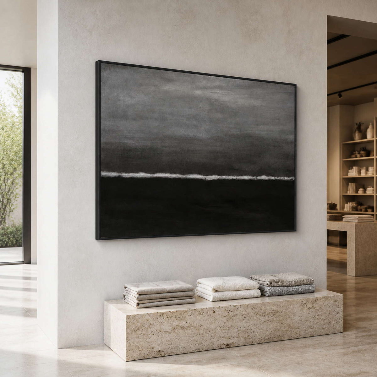 Black framed abstract wave painting above folded textiles in a natural light retail gallery.