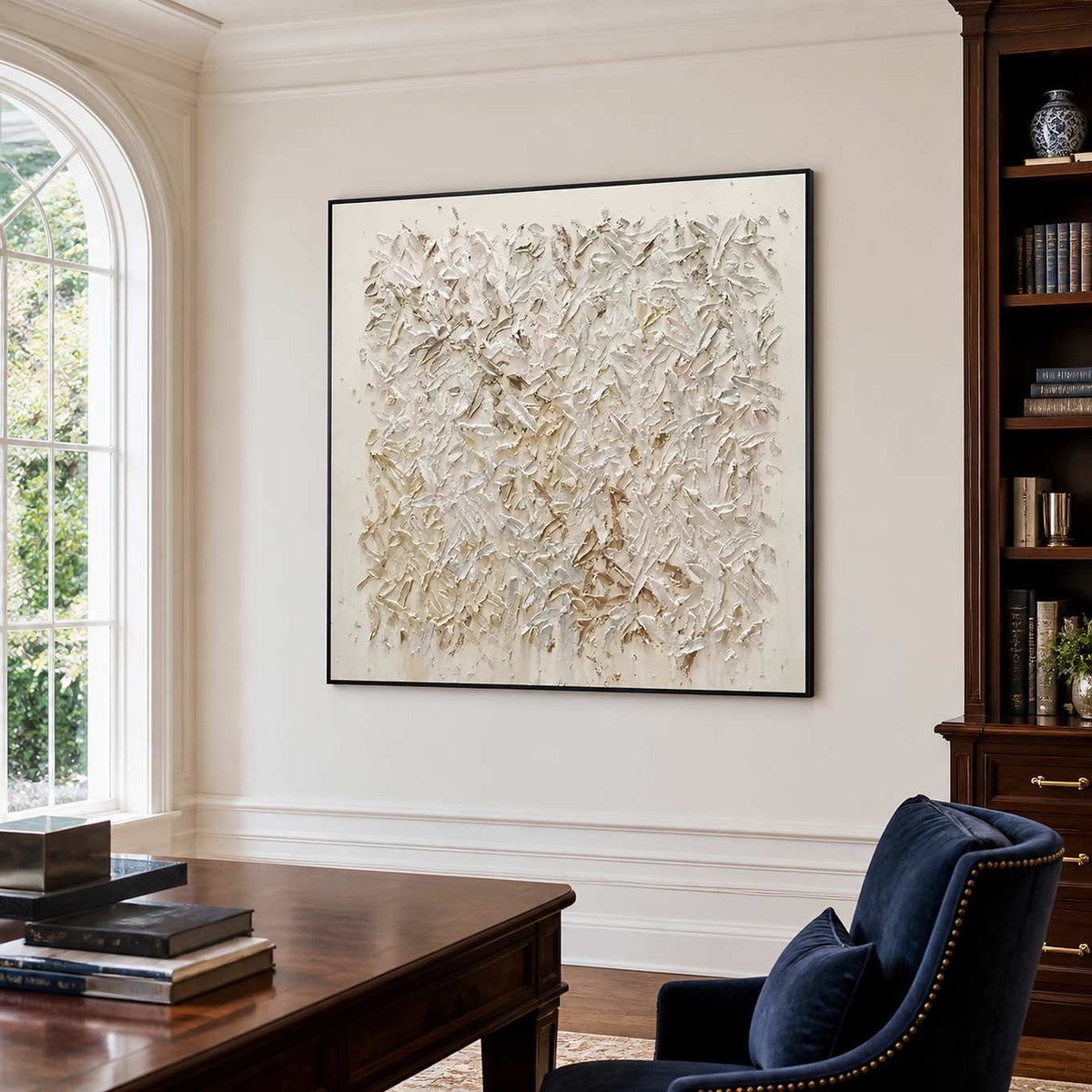 Black-framed white textured artwork beside a wooden desk and bookcase in a sunlit office.