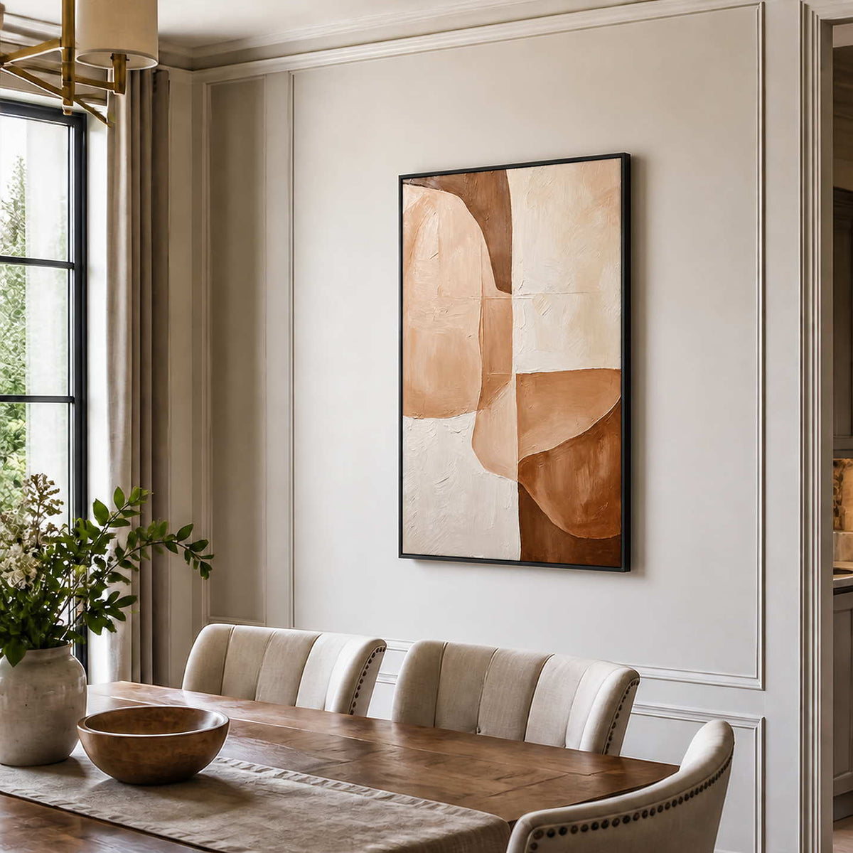 Black-framed earth-tone abstract painting above cream dining chairs beside a large window and wooden table.