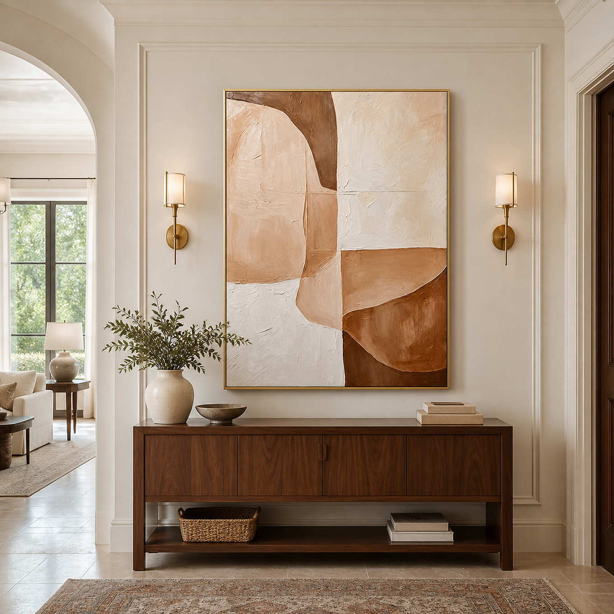 Gold-framed cream and caramel painting between wall sconces above a wooden hallway console.
