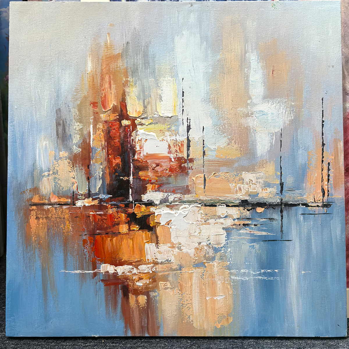 Photographed view of amber and rust blocks crossing a dark horizontal line on pale blue, with the artwork edge and surrounding background visible.