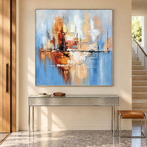 Amber and rust blocks crossing a dark horizontal line on pale blue in an artwork with a silver-colored frame, displayed in an entryway above a console or bench.