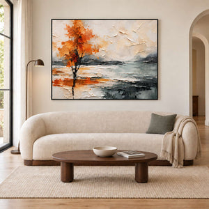 An orange autumn tree reflected beside a gray lake in a black frame, displayed above a sofa in a living room.