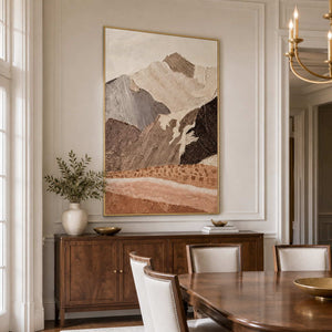 Gold framed textured mountain artwork above a wooden sideboard beside a dining table.