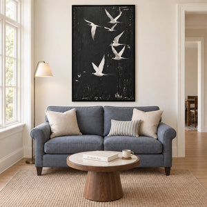 White birds flying across a charcoal-black sky in a black frame, displayed above a sofa in a living room.
