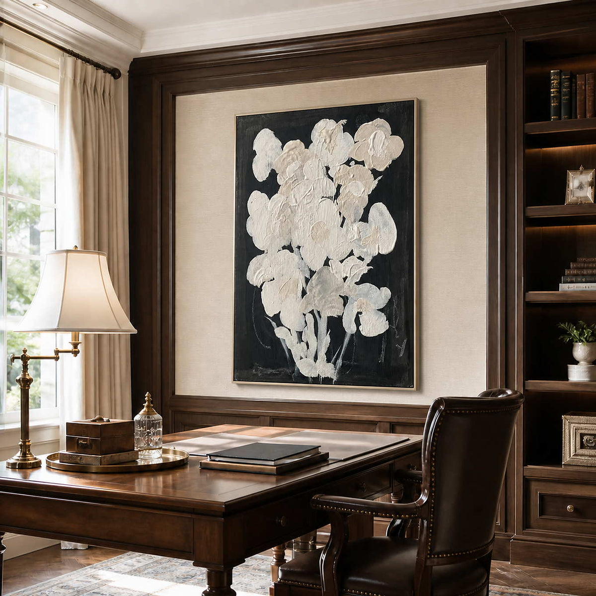 Silver framed cream floral artwork above a wooden desk between dark bookshelves.