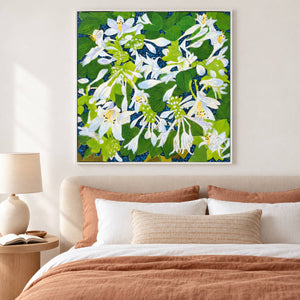 White flowers and broad green leaves over a deep blue ground in an artwork with a white frame, displayed in a bedroom near the bed.