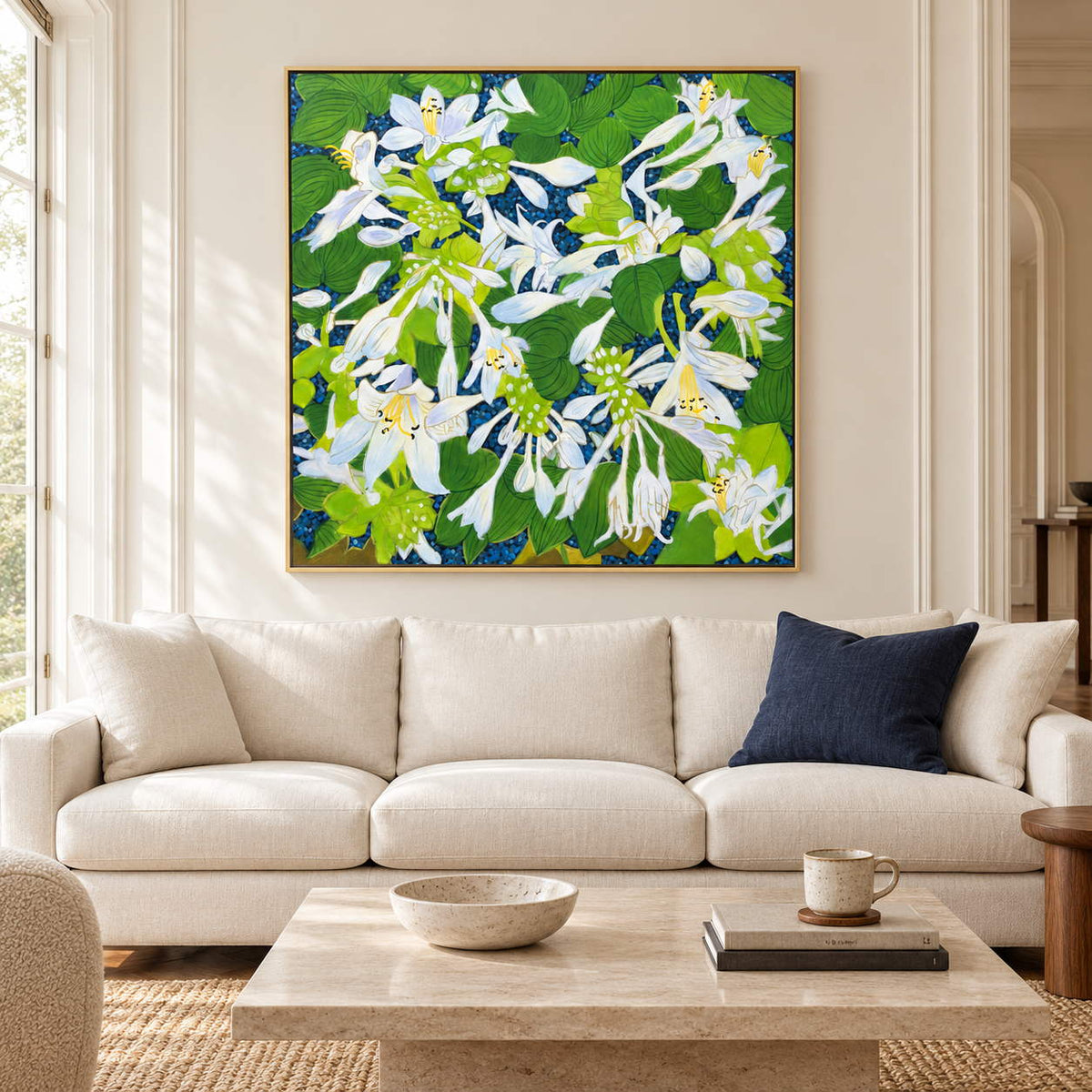 In a living room above seating, an artwork with a gold-colored frame shows white flowers and broad green leaves over a deep blue ground.