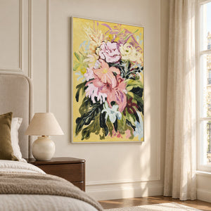 Large pink and cream flowers with dark green leaves on a warm yellow ground in an artwork with a white frame, displayed in a bedroom near the bed.