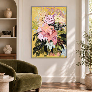 Artwork showing large pink and cream flowers with dark green leaves on a warm yellow ground beside a reading chair, with a silver-colored frame.