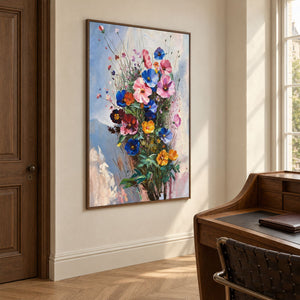 In a study with a desk, an artwork with a brown wood-look frame shows pink, blue and orange flowers on fine stems with dark green leaves against pale blue.