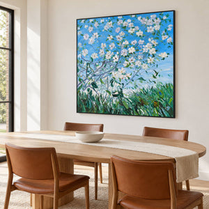 White flowering branches against blue sky above green grass in an artwork with a black frame, displayed in a dining area beside the table.