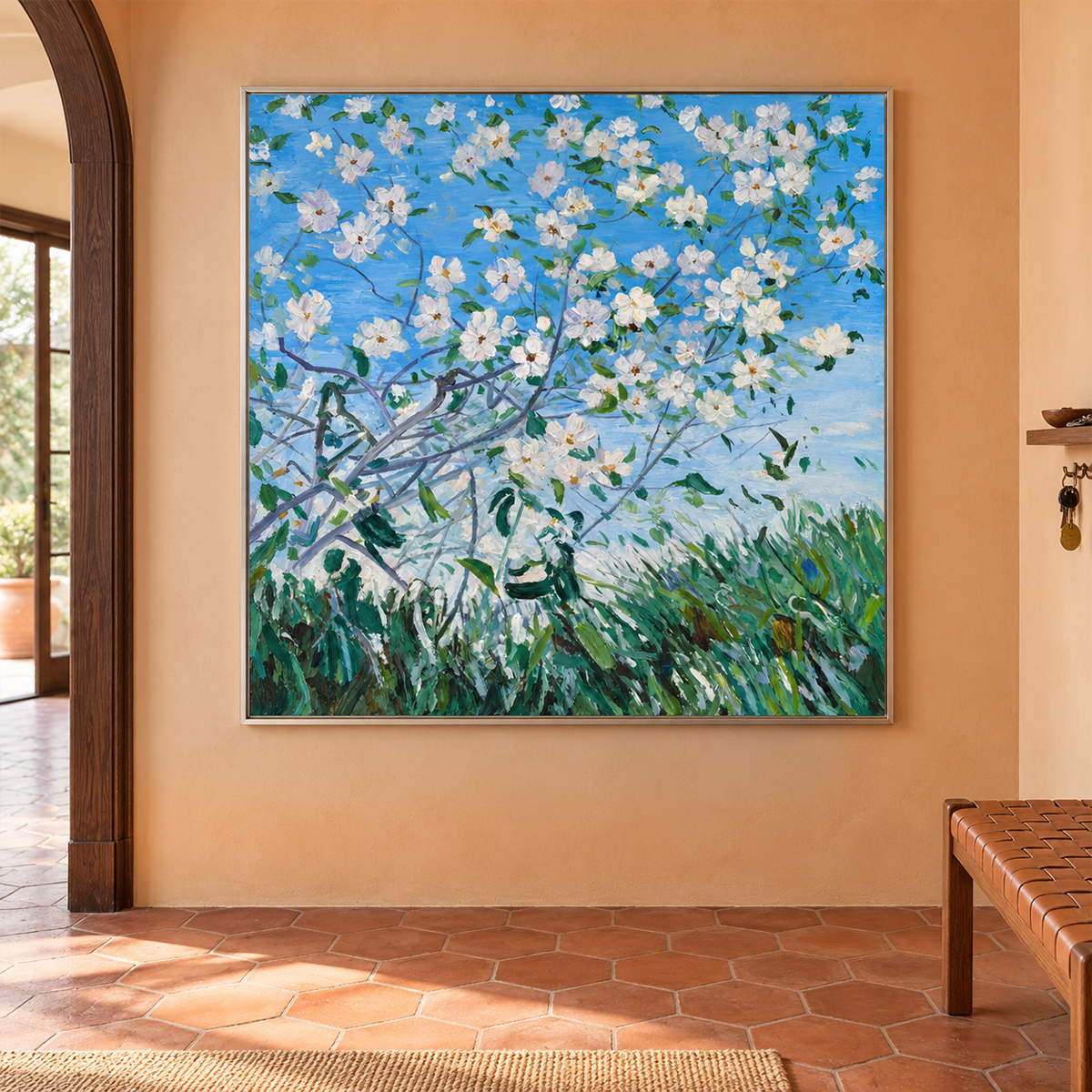 In an entryway above a console or bench, an artwork with a silver-colored frame shows white flowering branches against blue sky above green grass.