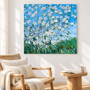 White flowering branches against blue sky above green grass in an artwork with no outer frame, displayed beside a reading chair.