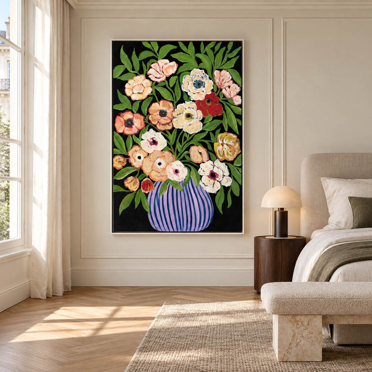In a bedroom near the bed, an artwork with a white frame shows cream, peach and red flowers with green leaves in a purple-blue striped vase on black.