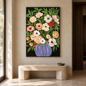 In an entryway above a console or bench, an artwork with no outer frame shows cream, peach and red flowers with green leaves in a purple-blue striped vase on black.