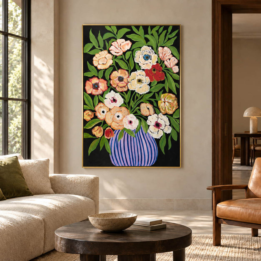 Cream, peach and red flowers with green leaves in a purple-blue striped vase on black in an artwork with a gold-colored frame, displayed in a living room above seating.