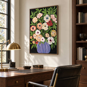Cream, peach and red flowers with green leaves in a purple-blue striped vase on black in an artwork with a brown wood-look frame, displayed in a study with a desk.