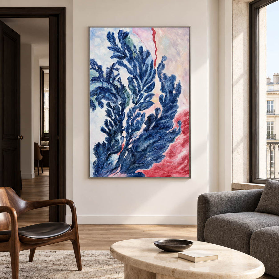In a living room above seating, an artwork with a silver-colored frame shows branching deep blue forms curling upward over pale pink, cream and red.