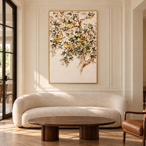 Branching gray-green leaves and pale yellow-green fruit on cream in an artwork with a gold-colored frame, displayed in a living room above seating.