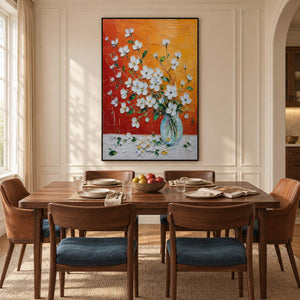 White blossoms in a glass vase against an orange and red background in a black frame in a dining room.