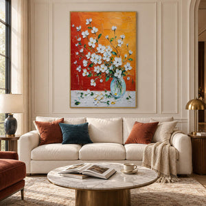 White blossoms in a glass vase against an orange and red background in a gold frame in a living room.