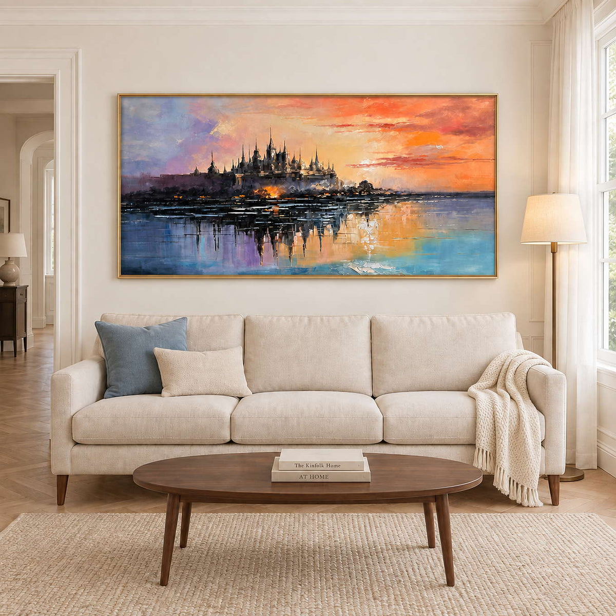 A distant castle reflected in water beneath an orange sunset in a gold frame, displayed above a sofa in a living room.