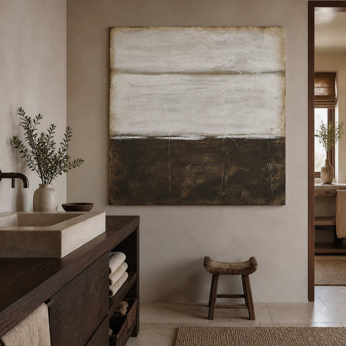 Frameless earth-toned abstract beside a rectangular stone bathroom basin and woven wooden stool.