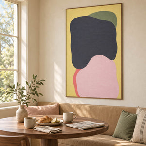 Yellow abstract painting with charcoal, blush, olive, and coral shapes in a brown wood frame in a breakfast dining area.