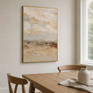 Gold framed pale landscape painting with creamy texture beside a wooden dining table.