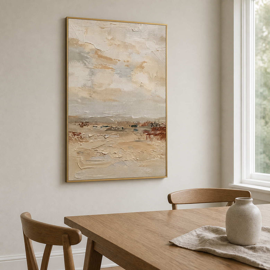 Gold framed pale landscape painting with creamy texture beside a wooden dining table.
