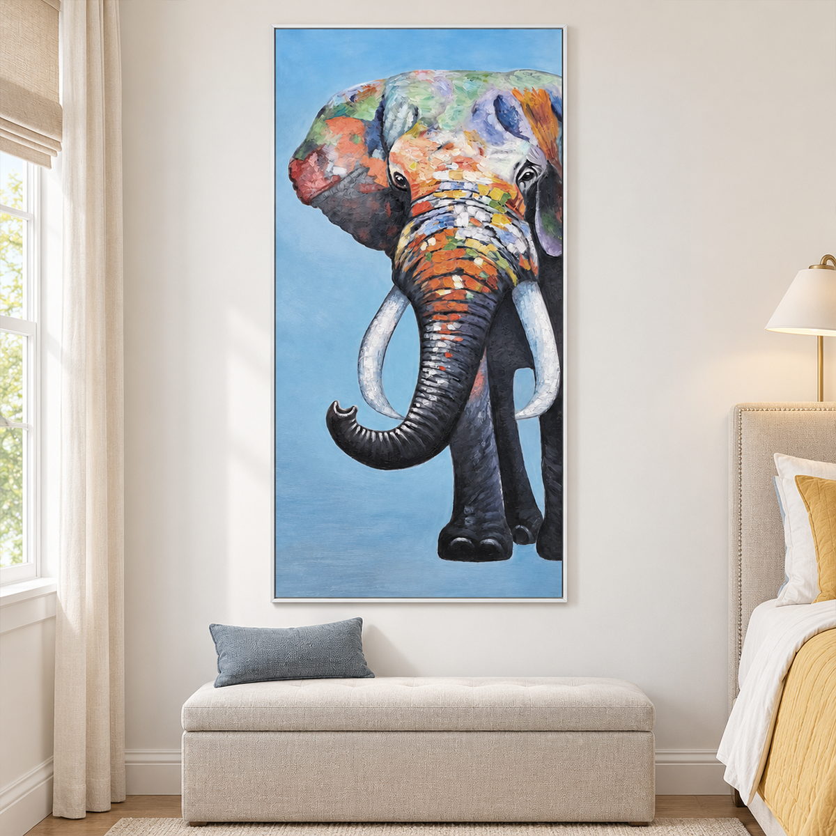 In a bedroom near the bed, an artwork with a white frame shows a front-facing elephant with an upcurled trunk, pale tusks and colored blocks on blue.