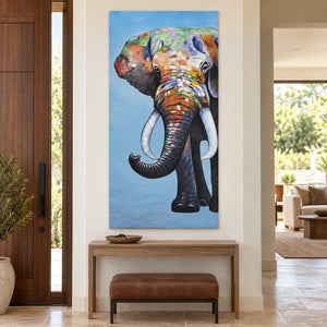 A front-facing elephant with an upcurled trunk, pale tusks and colored blocks on blue in an artwork with no outer frame, displayed in an entryway above a console or bench.