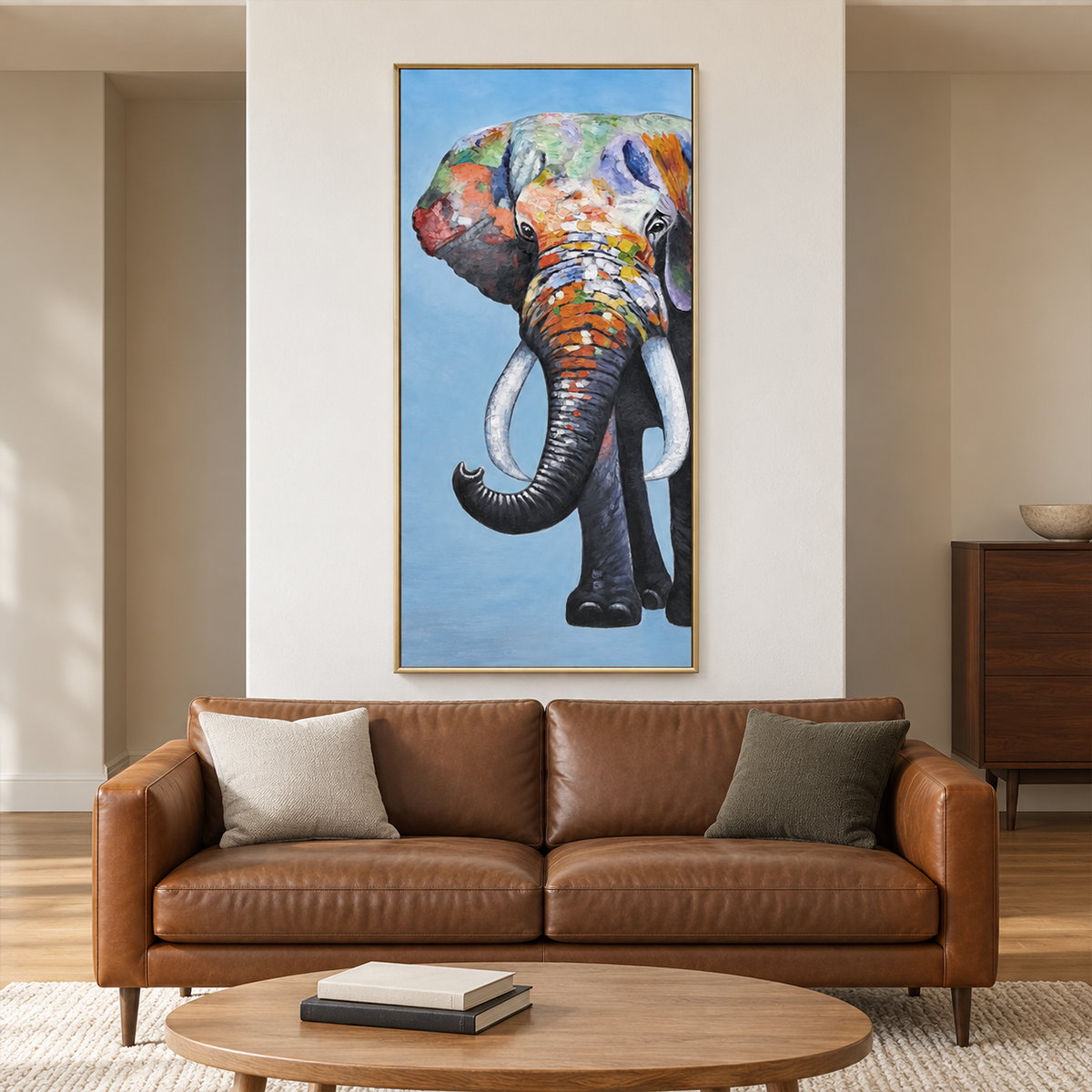 Artwork showing a front-facing elephant with an upcurled trunk, pale tusks and colored blocks on blue in a living room above seating, with a gold-colored frame.