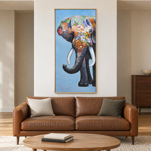 Artwork showing a front-facing elephant with an upcurled trunk, pale tusks and colored blocks on blue in a living room above seating, with a gold-colored frame.