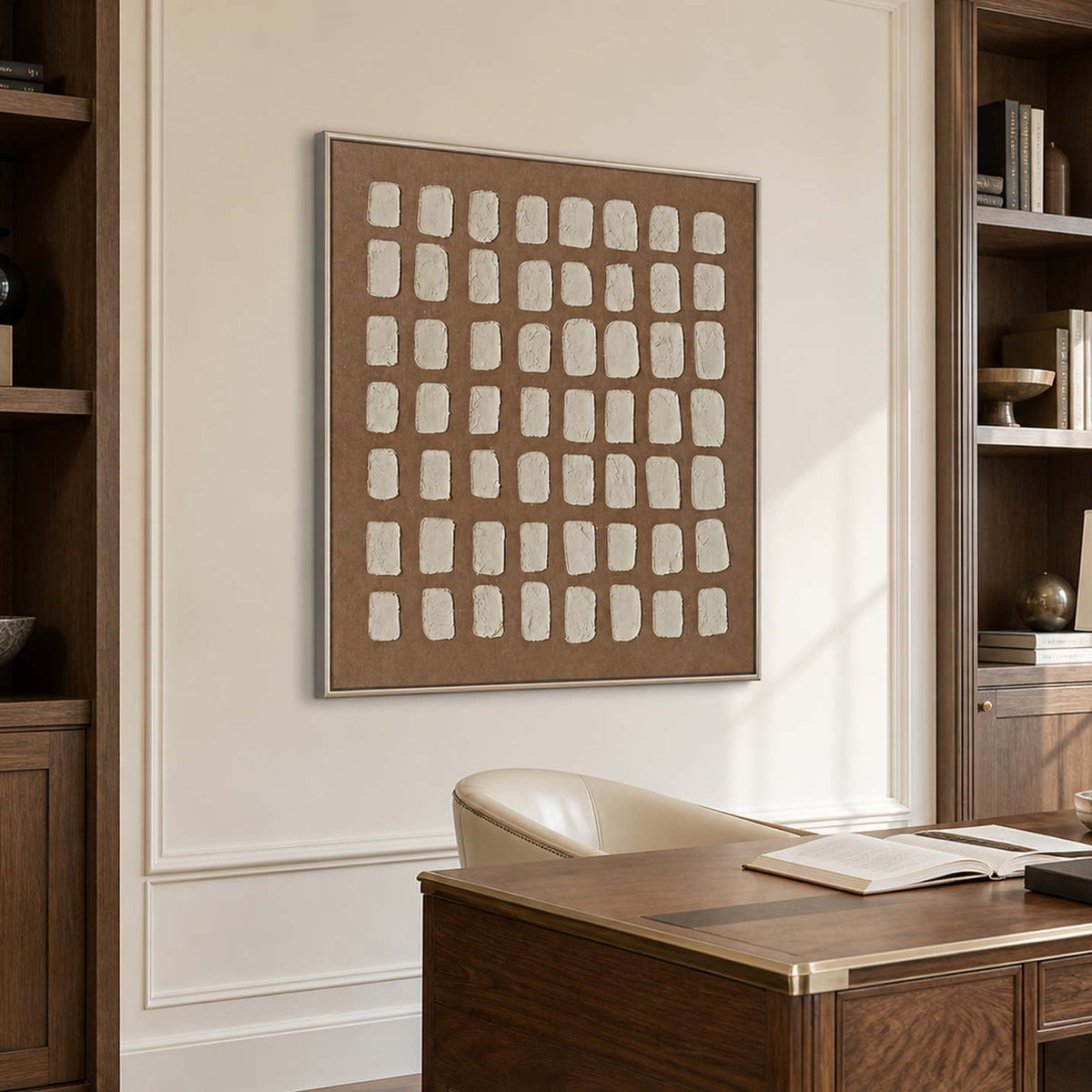 Silver framed cream grid artwork above a wooden desk between dark bookshelves.
