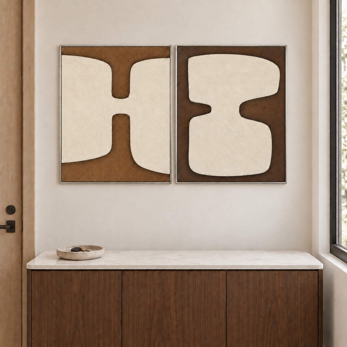 Two panels of complementary ivory organic forms on brown grounds in silver frames, displayed above an entryway console.