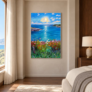Artwork showing a sunlit blue sea between low shores with colorful flowers in the foreground in a bedroom near the bed, with a white frame.