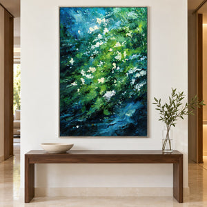 Artwork showing small white flowers among diagonal layers of green and deep blue strokes in a hallway above a bench, with a silver-colored frame.