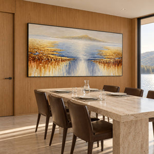 Artwork showing pale distant mountains above blue-gray water and gold-ochre marks spreading from both sides in a dining area beside the table, with a black frame.