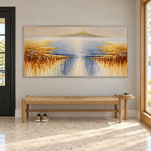 Artwork showing pale distant mountains above blue-gray water and gold-ochre marks spreading from both sides in an entryway above a console or bench, with a silver-colored frame.