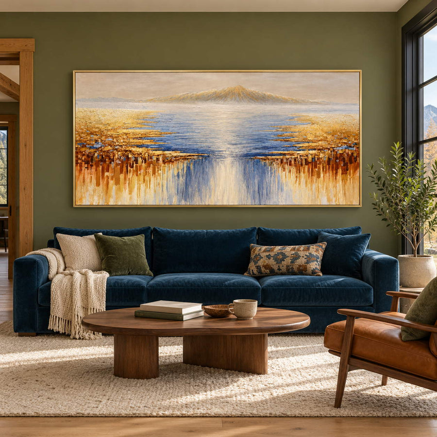 In a living room above seating, an artwork with a gold-colored frame shows pale distant mountains above blue-gray water and gold-ochre marks spreading from both sides.