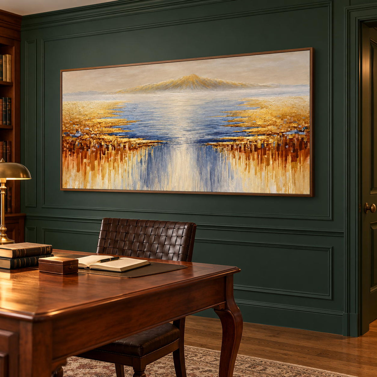 In a study with a desk, an artwork with a brown wood-look frame shows pale distant mountains above blue-gray water and gold-ochre marks spreading from both sides.