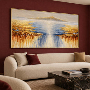 Pale distant mountains above blue-gray water and gold-ochre marks spreading from both sides in an artwork with a gold-colored frame, displayed in a seating area.