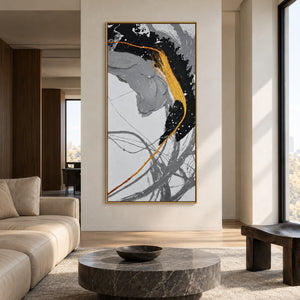 Broad black and gray brush arcs crossed by a gold-colored line and white flecks in an artwork with a gold-colored frame, displayed on a living-room wall beside a sofa.