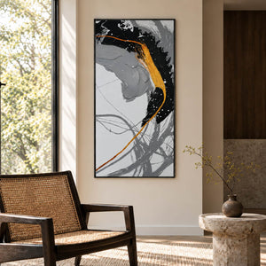 Broad black and gray brush arcs crossed by a gold-colored line and white flecks in an artwork with a black frame, displayed beside a reading chair.
