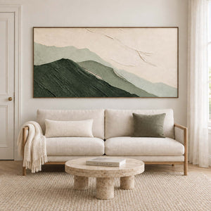 Layered dark and pale green mountains beneath an ivory sky in a brown wood frame, displayed above a sofa in a living room.