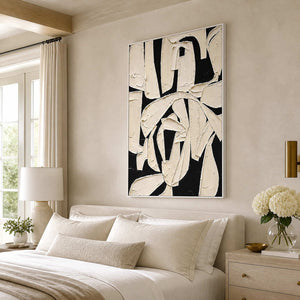 White framed ivory and black abstract painting above a pale bedroom headboard.