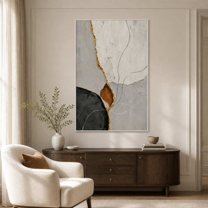 White framed abstract painting with a gold-toned seam above a dresser beside an upholstered chair.