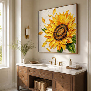 Wood-framed sunflower painting above a cream stone bathroom vanity with brass fittings and folded towels.